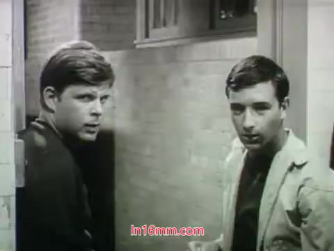 The still from the 1968 trailer for 'Out of It' features two young men, possibly in a hallway or corridor setting. They appear engaged in conversation, with a sense of curiosity or concern on their faces. The black-and-white format adds a classic feel to the scene, complementing the film's vintage aesthetic. Overall, it captures an intimate moment that hints at the storyline of the film.