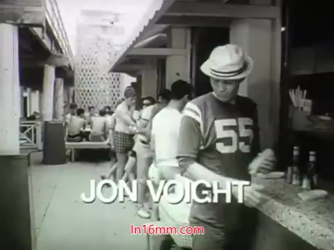 The still from 'Out of It' (1968) features a scene set in a casual outdoor setting, likely a cafe or diner. It prominently displays Jon Voight, who appears in a sports jersey with the number 55. The background shows people mingling and enjoying their time, capturing the relaxed vibe of the era. The overall aesthetic reflects the black-and-white film style typical of that period, enhancing its nostalgic charm.