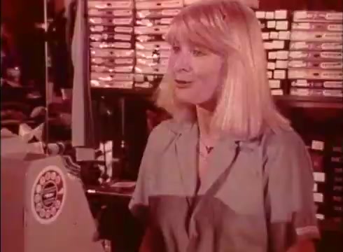 The still from the 'Telecheque commercial - circa 1980s' features a woman with blonde hair, wearing a light blue shirt, speaking to a customer. The backdrop shows shelves filled with products, indicating a retail environment. The conversation highlights the Telecheque service allowing customers to use checks confidently, emphasizing the ease and security it offers.