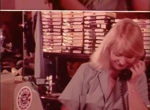 The 16mm still from the 'Telecheque' commercial features a woman in a light blue shirt, smiling as she speaks on the phone. She appears engaged in a conversation about payment by check, showcasing the Telecheque service as a means to ensure checks are valid. In the background, tape or storage boxes are visible, suggesting a business setting. The overall tone is friendly and reassuring, typical of commercial messaging from the 1980s.