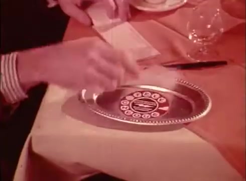 The still depicts a scene from a 1980s Canadian French commercial for the Telecheque service. A hand is seen placing a check on a silver platter, possibly in a restaurant setting. The background includes a table with a tablecloth and dining utensils. The dialogue suggests a conversation around using checks for payment, highlighting the convenience of the Telecheque service for verifying check validity.