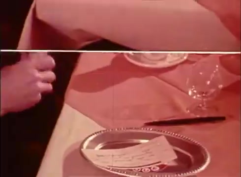 The 16mm still from the 'Telecheque commercial - circa 1980s' features a close-up of a dining table setting. A person’s hand is seen handling a piece of paper, likely a check, with a silver plate beneath it. The ambiance suggests a casual dining experience, and the colors are warm and slightly faded, typical of the era's film quality. The transcription highlights a conversation about using a check for payment, emphasizing the Telecheque service's convenience.