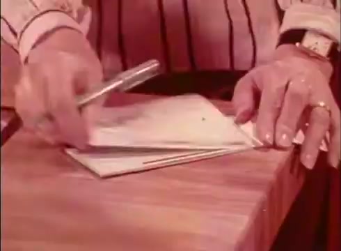 The still from the Telecheque commercial features a person's hands writing on a check at a wooden counter. The individual is dressed in a striped shirt, and there is a pen in one hand while they grip the check with the other. The scene conveys a sense of everyday transactions and emphasizes the convenience of the Telecheque service being discussed in the dialogue, which centers around payment by check and the assurance it provides.