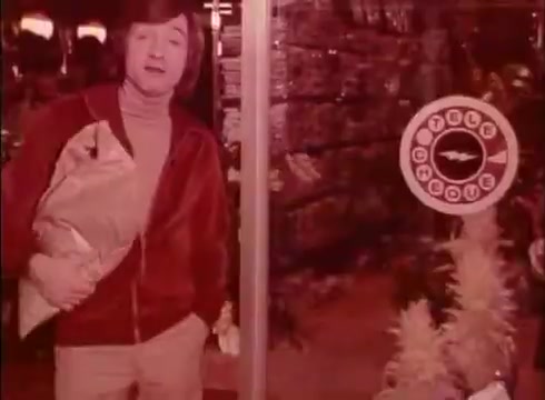 The still from the 'Telecheque commercial - circa 1980's' features a man dressed in a red tracksuit, standing in front of a store window. He is holding a package and looks directly at the camera. Behind him, there is a circular sign with the logo for Telecheque, emphasizing the service that enables businesses to verify checks before accepting them. The overall tone is casual and friendly, typical of advertisements from that era. The commercial conveys a sense of convenience and assurance regarding check payments.