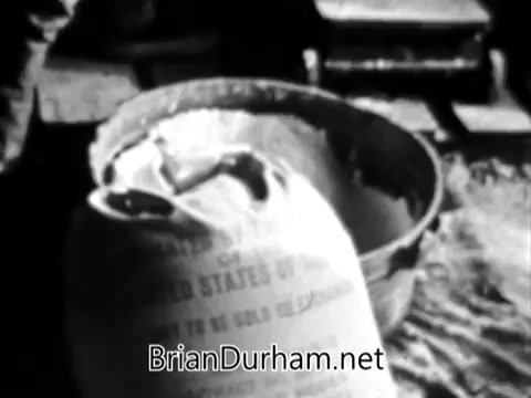 The still from the 'Catholic Bishops' Overseas Aid Fund PSA' depicts a scene emphasizing the disparity in resources between affluent areas and those suffering from poverty. It shows a bag labeled as food aid, likely from the United States, placed next to a metal container, suggesting efforts to provide sustenance to those in need. The imagery underscores the urgent call to action to support humanitarian relief efforts and address issues like high infant mortality and malnutrition in impoverished regions. The overall tone is one of solidarity and a plea to help close the widening gap between abundance and scarcity.