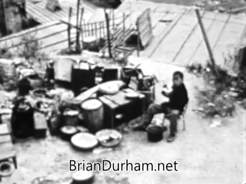 The still features a stark, barren environment filled with various containers and debris, indicative of poverty and hardship. In the foreground, a lone figure sits, possibly engaged in a menial task, symbolizing struggle and resilience amidst challenging conditions. This imagery serves to highlight the disparity between abundance and scarcity, aligning with the message of the Catholic Bishops' Overseas Aid Fund PSA about the growing gap in resources and the urgent need for support to those in need.
