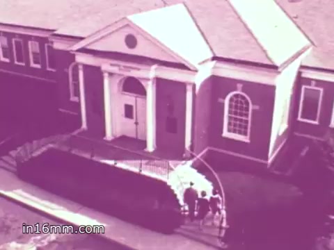 The still depicts a historic building, presumably the American Freedom Center, characterized by its classical architectural elements, such as columns and large windows. There are a few figures walking toward the entrance, set against a muted color palette. The context involves a voiceover by Anita Bryant referencing the struggles of George Washington’s army during the harsh winter at Valley Forge, reinforcing themes of sacrifice and freedom in America. The overall tone is nostalgic and patriotic.