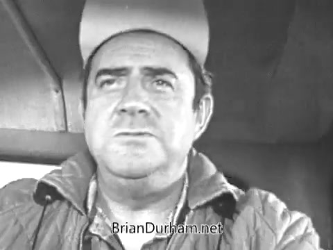 The still features a man with a serious expression, wearing a cap and a jacket, seated presumably in a vehicle. The black-and-white image captures a moment that may convey contemplation or focus, reflective of the advertising style of the era. It likely emphasizes authenticity and connection to everyday life, consistent with the themes in commercials of the late 1960s.