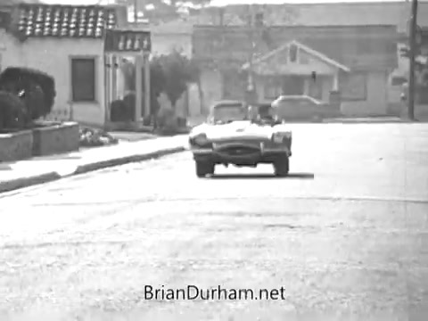 The still from the 'Red Rose Tea' commercial features a street scene with a vintage convertible car driving down a quiet road. The environment has mid-20th-century architecture, including low buildings and manicured landscaping. The overall atmosphere suggests a nostalgic feel, typical of commercials from that era. The focus on the car and streetscape likely evokes themes of leisurely moments and enjoyment.