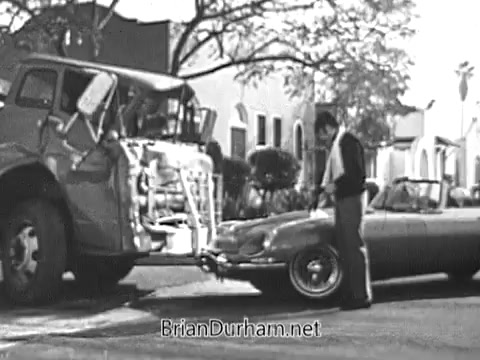 The still from the 1968 Red Rose Tea commercial showcases a scene where a man is interacting with a vintage car parked next to a truck. The setting appears to be a street lined with trees and buildings in the background. The focus is on the man, who is dressed casually, suggesting a lighthearted and relatable moment, characteristic of commercial narratives from that era. The overall tone is nostalgic and captures the charm of the 1960s, emphasizing everyday life.