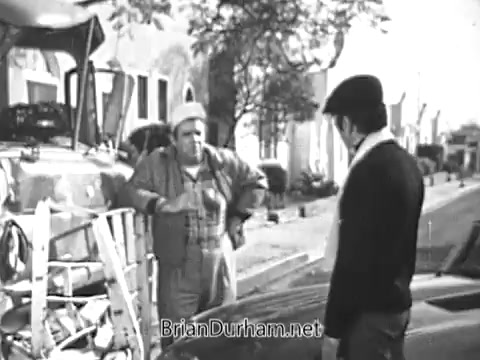 The still from the 1968 Red Rose Tea commercial features two characters engaging in a conversation outdoors. One character, dressed casually with a hat, appears to be listening intently to the other, who is wearing a work outfit and a cap. The setting includes a vehicle with visible tea branding in the background, surrounded by trees and buildings typical of a neighborhood scene. The overall atmosphere suggests a friendly exchange, likely related to the benefits or enjoyment of drinking tea.