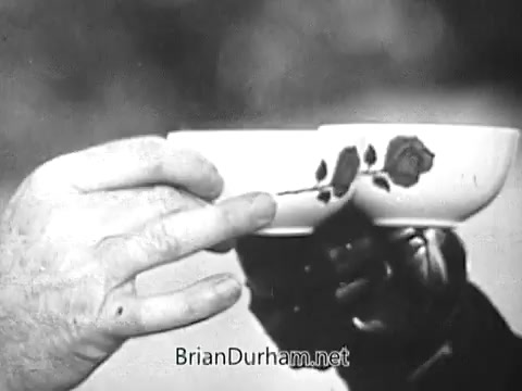 The still from the 1968 Red Rose Tea commercial features hands holding two delicate teacups, each adorned with a rose design. The focus on the cups suggests a moment of sharing or camaraderie, typical of tea culture. The scene conveys a warm, inviting atmosphere associated with the beverage.