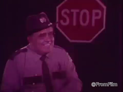 The still showcases a smiling man in a police uniform, possibly a traffic officer, standing in front of a stop sign. The lighting appears to have a pinkish hue, typical of some promotional materials from the 1970s. The context focuses on promoting the use of reflector bulbs to save electricity, likely presenting a friendly and approachable demeanor to convey the message effectively.