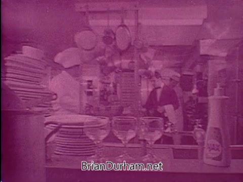 The still from the Ajax Dishwashing Liquid commercial features a kitchen setting, with chefs and staff actively involved in dishwashing. The scene shows an array of stacked plates and glassware, emphasizing the cleaning process. The image has a pinkish hue, likely due to the film quality or processing. The focal point appears to be Ajax Dishwashing Liquid, positioned prominently as part of the brand's promotion.