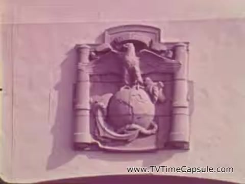 The still features an emblematic insignia of the Marine Corps, prominently displaying an eagle, globe, and anchor. The design is set against a light-colored background, indicative of military architecture. This image reflects the values of strength and commitment, aligning with the themes of the Marine Corps recruiting spot from the Vietnam era, which aimed to portray the building of character and resilience among recruits.