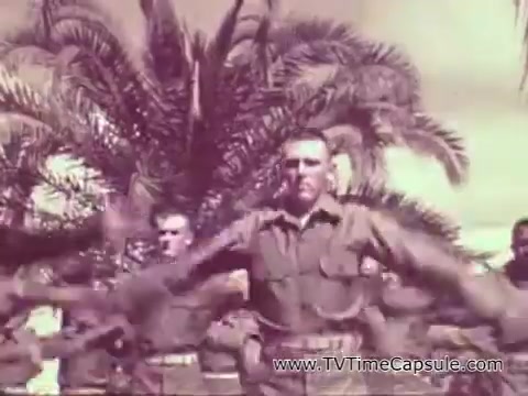 The still from 'Marine Corps Recruiting Spot - The Marine Corps Builds Men' depicts a group of Marines in uniform engaged in synchronized physical training under a palm tree backdrop. The image highlights military discipline and camaraderie, reflective of the Vietnam era's recruitment efforts emphasizing strength and teamwork. The overall tone conveys a sense of pride and dedication to service.