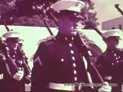The still depicts a group of Marines in formal uniform, likely during a ceremonial event. The soldiers are standing at attention, showcasing a disciplined military presence. The image reflects the ideals of strength and unity emphasized in Marine Corps recruiting efforts during the Vietnam era. The color tone suggests a vintage aesthetic, typical of 16mm film from that time period.