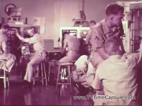 The still from the Marine Corps recruiting spot depicts a group of young men in military uniforms engaged in various activities. The scene suggests a training or educational environment, likely focused on teamwork and discipline, emblematic of the Marine Corps ethos during the Vietnam era. The setting appears to be a classroom or workshop, with equipment and chairs, showcasing a hands-on approach to building skills and camaraderie. Overall, the image conveys the message of the Marine Corps as a transformative experience for young men.