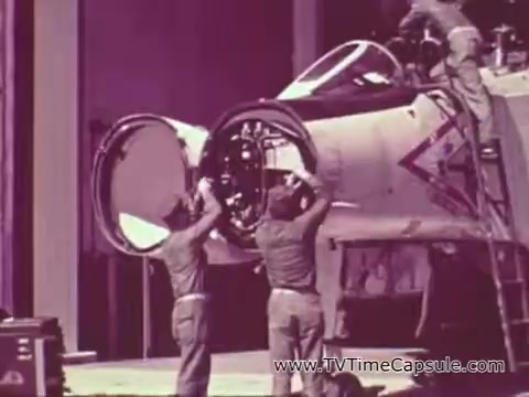 The still from the Marine Corps recruiting spot features two Marines working on an aircraft, likely in a hangar setting. The image captures the technical aspect of military training during the Vietnam era, emphasizing teamwork and skill development. The color tones suggest a vintage look, characteristic of the period's film quality. The focus is on the Marines' dedication to their craft, aligning with the theme of building men through rigorous training and discipline.
