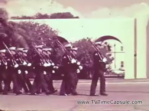 The still captures a group of uniformed Marine Corps recruits marching in formation with rifles, likely during a training or ceremonial event. The setting appears formal, with a backdrop of a building that suggests a military installation. The image reflects the disciplined nature of military training during the Vietnam era, emphasizing themes of service, camaraderie, and the recruitment of young individuals into the Marine Corps. The color palette and visual style are characteristic of that period in film.