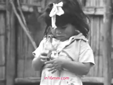 The still features a young girl with long hair, wearing a light-colored shirt. She appears to be looking down at her hands, engaged in a moment of quiet introspection. The background suggests a simple outdoor setting, possibly reflective of the 1960s. The image conveys a sense of innocence and contemplation, fitting the theme of a public service announcement aimed at fostering awareness and compassion.