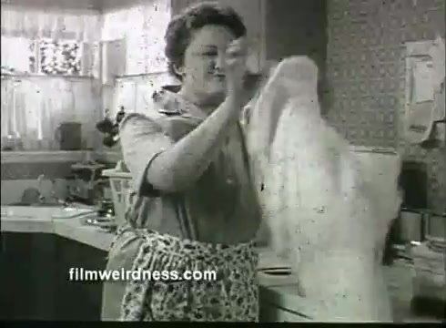 The still features a woman in a vintage kitchen from the 1960s, engaged in the process of laundry. She is wearing a light-colored dress and an apron, looking focused as she holds a white garment. The kitchen has classic elements of the era, including patterned wallpaper and a window with sheer curtains, reflecting the domestic theme typical of advertisements from that time. The scene conveys a sense of homemaking and the use of Ajax Laundry Detergent.