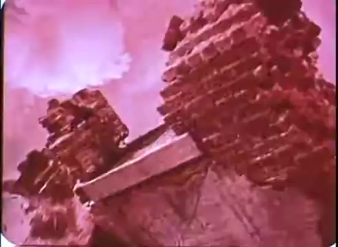 The still from 'The Time Machine' promo showcases a desolate landscape featuring crumbling brick structures. The image has a reddish tint, suggesting a post-apocalyptic or futuristic setting. Dramatic clouds loom in the background, enhancing the eerie atmosphere of decay and time. The overall composition conveys a sense of abandonment and the passage of time.