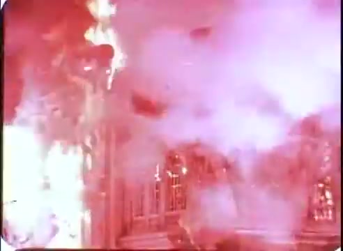 The still from the promo for 'The Time Machine' depicts a dramatic scene of destruction, characterized by bright flames and dense smoke. The intense red and orange hues suggest a sense of chaos and urgency, possibly indicating a catastrophic event within the narrative. Visual elements like the blurred outlines of the environment hint at a larger setting being engulfed in fire, effectively conveying the film's themes of time and consequence.