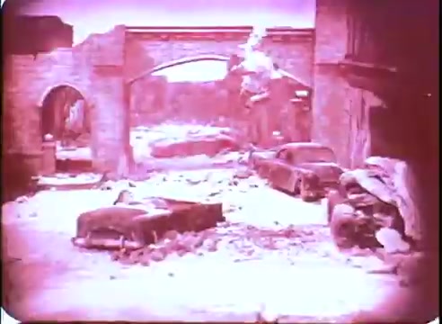 The still from 'The Time Machine' promo depicts a desolate urban landscape, characterized by crumbling architecture and abandoned vehicles. The scene has a pinkish tint, enhancing the post-apocalyptic atmosphere. Debris litters the ground, suggesting devastation and neglect, while the arches in the background hint at a once-thriving city now left in ruins. The overall mood evokes a sense of abandonment and the passage of time.