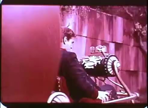 The still from 'The Time Machine' promotional material shows a man seated in a vintage time travel machine, which is intricately designed with mechanical details. The color tone has a reddish hue, typical of 16mm prints, and the background features a blurred, abstract wall, enhancing the focus on the character and the machine. The overall composition suggests a moment of anticipation or adventure.
