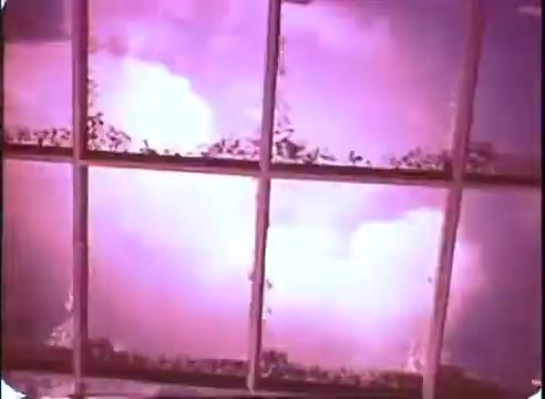 The still from 'The Time Machine' promo features a window with a grid pattern, revealing a dramatic sky filled with clouds. The colors are predominantly pink and purple, creating a surreal and atmospheric effect. The scene conveys a sense of wonder and intrigue, fitting the film's themes of time travel and adventure. The 16mm print adds a vintage quality to the image.