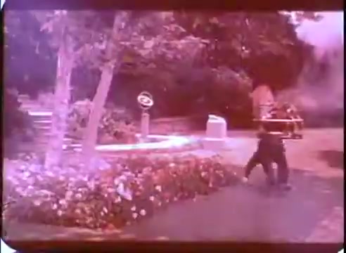 The still captures a scene from 'The Time Machine' promos, featuring a character in a garden setting with vibrant flowers and trees. The image exhibits a hazy, dreamy quality typical of 16mm prints, with a notable color palette. A vintage-looking device is partially visible, hinting at the film's science fiction elements, while the character appears to be in motion, possibly reacting to an unseen event or phenomenon.