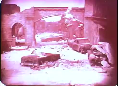 The still from 'The Time Machine' promotional material depicts a desolate urban landscape. Ruined buildings and debris dominate the scene, while abandoned, battered cars lie scattered across the ground. The atmosphere conveys a sense of decay and a post-apocalyptic world, enhanced by the film's distinctive color tones reminiscent of mid-20th century cinematography.