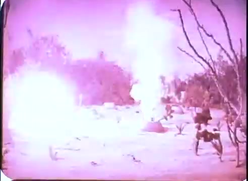 The 16mm still from the promos of 'The Time Machine' depicts a dramatic scene featuring explosions in a desert-like landscape. The image has a pinkish tint, highlighting the intensity of the blasts. Sparse vegetation and some leafless trees are visible in the background, lending a desolate atmosphere to the scene. Overall, it captures a moment of action and intrigue from the film.