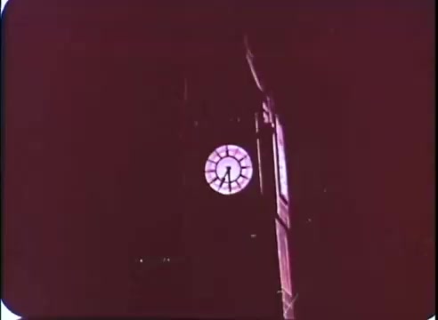 The still features a clock tower, prominently displaying a large clock face. The time is indicated at approximately 8 o'clock, with a warm, reddish tint dominating the image. The surrounding architecture suggests historical significance, evoking a sense of time and nostalgia, which aligns with the themes of 'The Time Machine.' The image captures an atmospheric moment, likely meant to convey the passage of time in the context of the film's narrative.