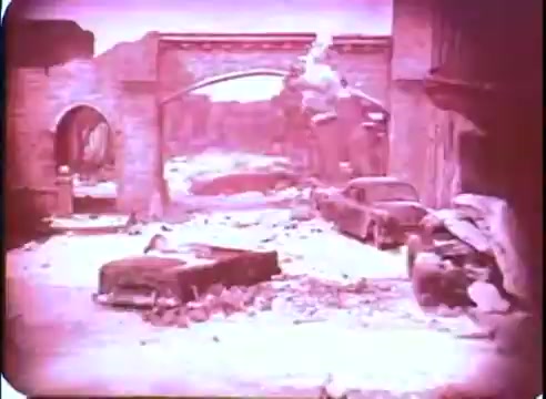 The still features a desolate urban landscape, characterized by crumbling structures and debris scattered throughout. An arched bridge spans the background, partially obscured by destruction. Abandoned cars are visible in the foreground, contributing to the post-apocalyptic atmosphere, with a muted color palette creating a sense of stark eerie desolation. The composition evokes themes of time and decay, aligning with the narrative of 'The Time Machine.'