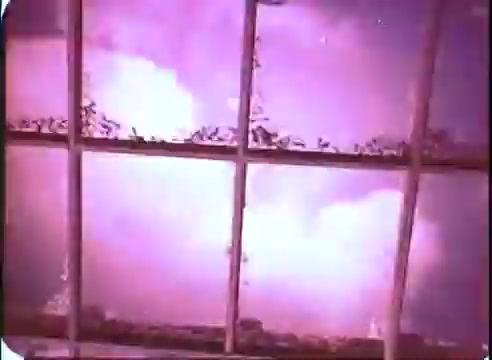 The still features a view through a gridded window. The scene is suffused with a purple hue, suggesting an otherworldly or surreal atmosphere. Wisps of cloud or smoke are visible beyond the glass, enhancing the sense of mystery or impending action, typical of sci-fi themes presented in promotional material for 'The Time Machine.' The aged quality of the 16mm print adds a nostalgic touch to the image.