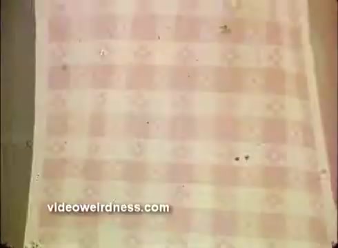 The still features a textured fabric with a pink and white checkered pattern, possibly representing a tablecloth. The backdrop evokes a festive atmosphere suitable for a Mardi Gras-themed setting, hinting at celebration and gatherings. The overall aesthetic is vintage, characteristic of a 16mm film quality.