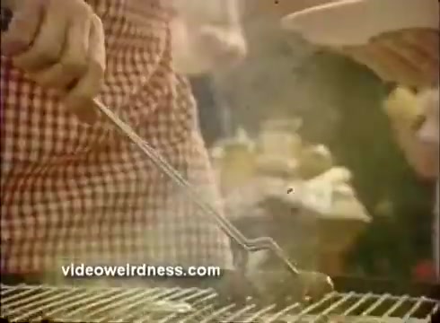 The still shows a close-up of a hand gripping a long-handled utensil, lifting food from a grill. The person is wearing a checked apron and surrounded by a light haze of steam, suggesting a festive cooking scene. The background implies a lively atmosphere, typical of a Mardi Gras celebration, highlighting the enjoyment of food in the context of a commercial for Mardi Gras paper products.