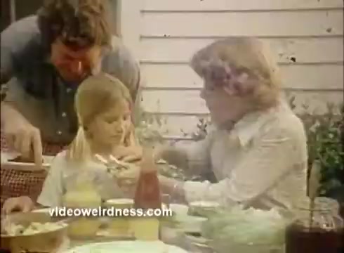 The still shows a family scene outdoors, focusing on a table set with various food items. A man serves food to a young girl, while a woman sits beside her, possibly assisting. The setting suggests a casual, friendly gathering, highlighting the use of Mardi Gras paper products, likely in the context of a festive meal or celebration. The overall tone is warm and inviting.