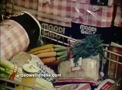 The still features a shopping cart filled with various grocery items, prominently displaying Mardi Gras paper products in the background. Notable items include carrots and other grocery essentials, surrounded by packaging that reinforces the festive theme associated with Mardi Gras celebrations. The overall imagery conveys a sense of celebration and preparation for festivities.