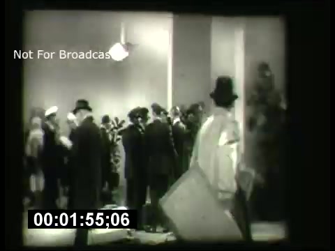 The still from 'ABC Sunday Night Movie promos - 1960s' depicts a scene in black and white, featuring a crowded setting with a group of people, some in hats and formal attire. A figure in a lighter coat appears to be moving through the scene, possibly carrying a large item. The overall atmosphere suggests a cinematic context, characteristic of the era's film promotions. The timestamp indicates a specific moment in the footage.
