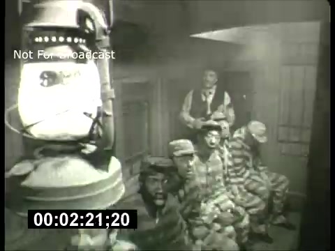 The still from the ABC Sunday Night Movie promos in the 1960s features a dimly lit scene with a group of men seated in what appears to be a jail or prison setting. They are wearing striped prison uniforms, conveying a tense or dramatic atmosphere. A lantern is positioned prominently in the foreground, giving the scene a nostalgic black-and-white aesthetic typical of that era. The composition suggests a moment of captivity or reflection among the characters, hinting at the themes often explored in films during that time.