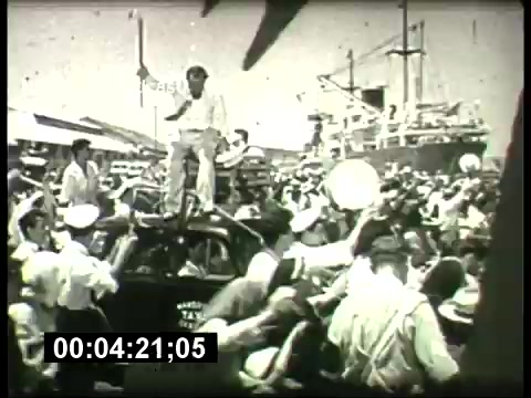 The still likely depicts a lively scene from a 1960s ABC Sunday Night Movie promo, featuring a bustling crowd around a car. A man stands on top of the vehicle, engaging with the crowd, while ships are visible in the background, suggesting a port setting. The image captures the energetic vibe typical of movie promotions from that era.