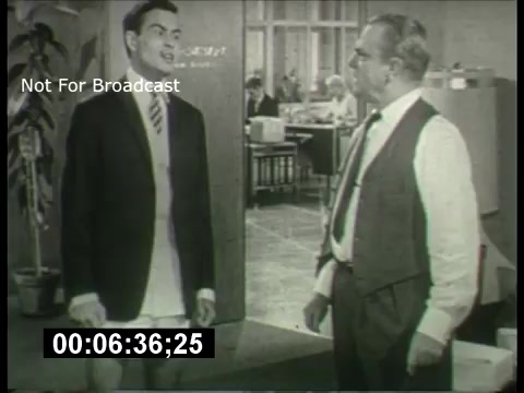The still features two characters engaged in a dialogue within an office setting. One character is dressed in a suit, presenting a formal appearance, while the other, slightly older, is in a vest and shirt, possibly representing authority or experience. The background includes desks and people working, signifying a bustling office environment typical of the 1960s. The black-and-white aesthetic emphasizes the era, and the timestamp indicates this is a still from a promotional segment for the ABC Sunday Night Movie. The 'Not For Broadcast' label suggests this content is intended for internal use or archival purposes.