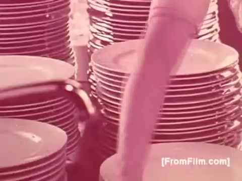 The still from 'Dove for Dishes' features a close-up of a woman's arm as she reaches for a stack of plates. The scene conveys a sense of mundane domesticity, with numerous plates neatly stacked in the background. The color palette suggests a pinkish hue, characteristic of the film's aesthetic from the 1970s. Overall, it captures a moment of everyday life, emphasizing domestic chores.