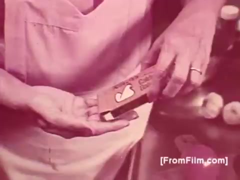 The still shows a person, likely a cook, holding a packet of Knorr Caldo De Galinha soup mix. The image has a warm, nostalgic tone typical of the 1970s, with a focus on the product. The background features a kitchen setting with ingredients, emphasizing home cooking. The cook's attire suggests a domestic environment, reinforcing the idea of family meals.