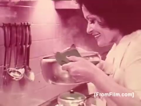 The still depicts a cheerful woman in a kitchen, holding a steaming pot. She smiles as she prepares a meal, likely featuring Knorr Caldo De Galinha soup. The environment is cozy, with kitchen utensils hanging in the background and rising steam adding warmth to the scene. The color palette suggests a retro aesthetic typical of 1970s advertisements.
