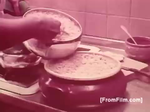 The still captures a kitchen scene from a 1970s Brazilian Knorr Caldo De Galinha soup commercial. A hand is seen pouring a mixture from a bowl into a pot, emphasizing the preparation of a hearty soup. The background features a simple kitchen setting, with a cozy, home-cooked atmosphere. The color tone suggests a vintage aesthetic typical of that era.