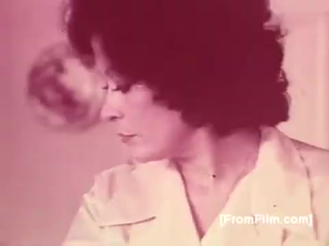 The still from the Knorr Caldo De Galinha soup commercial shows a woman with curly hair, looking to the side, engaged in a cooking activity. She appears focused and is dressed in a light-colored outfit, conveying a domestic setting typical of 1970s Brazil. The overall tone is warm, with a soft color palette, evoking a sense of comfort associated with home-cooked meals.